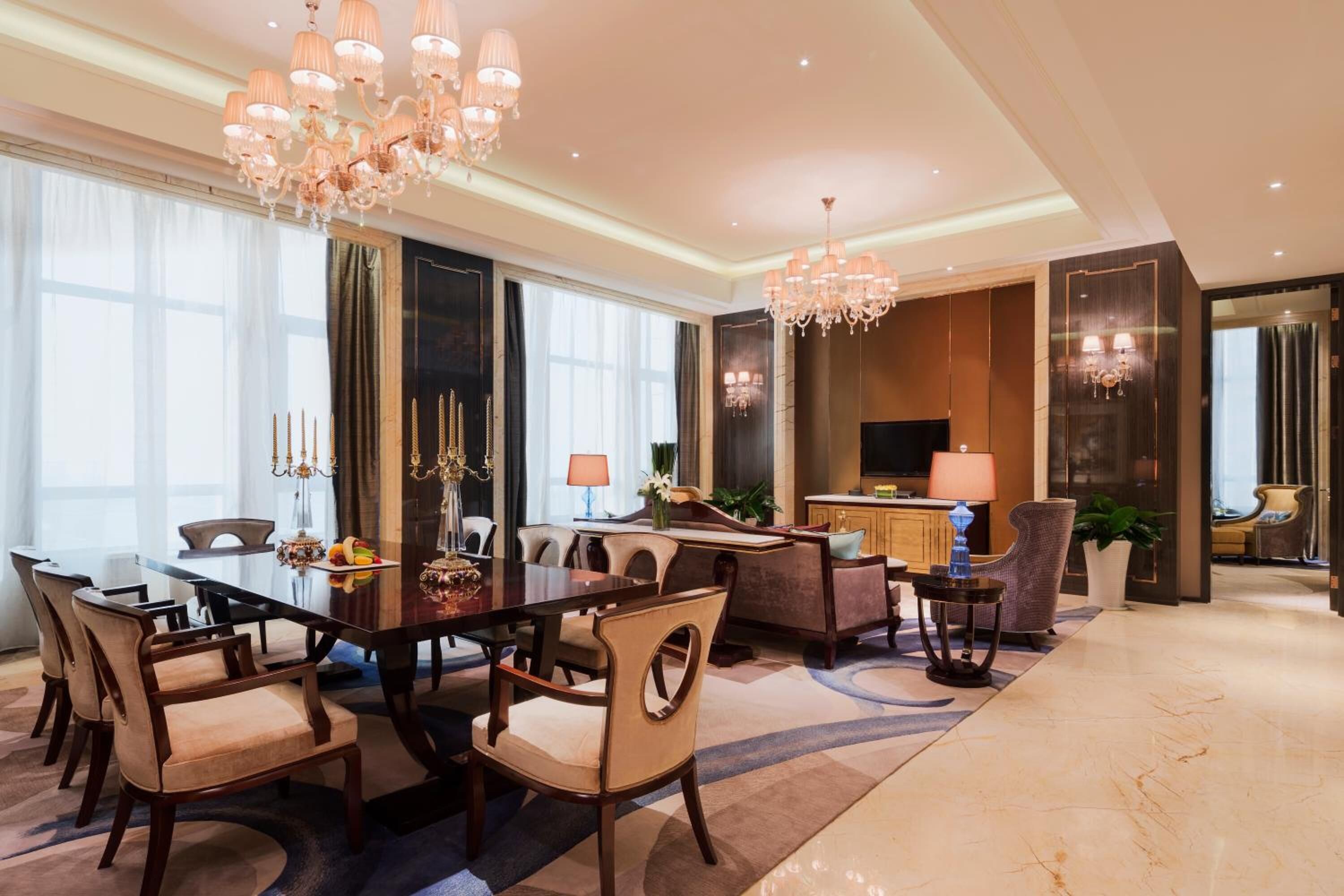 A spacious hotel room with a large dining table, comfortable seating, a television, and a well-lit living area.