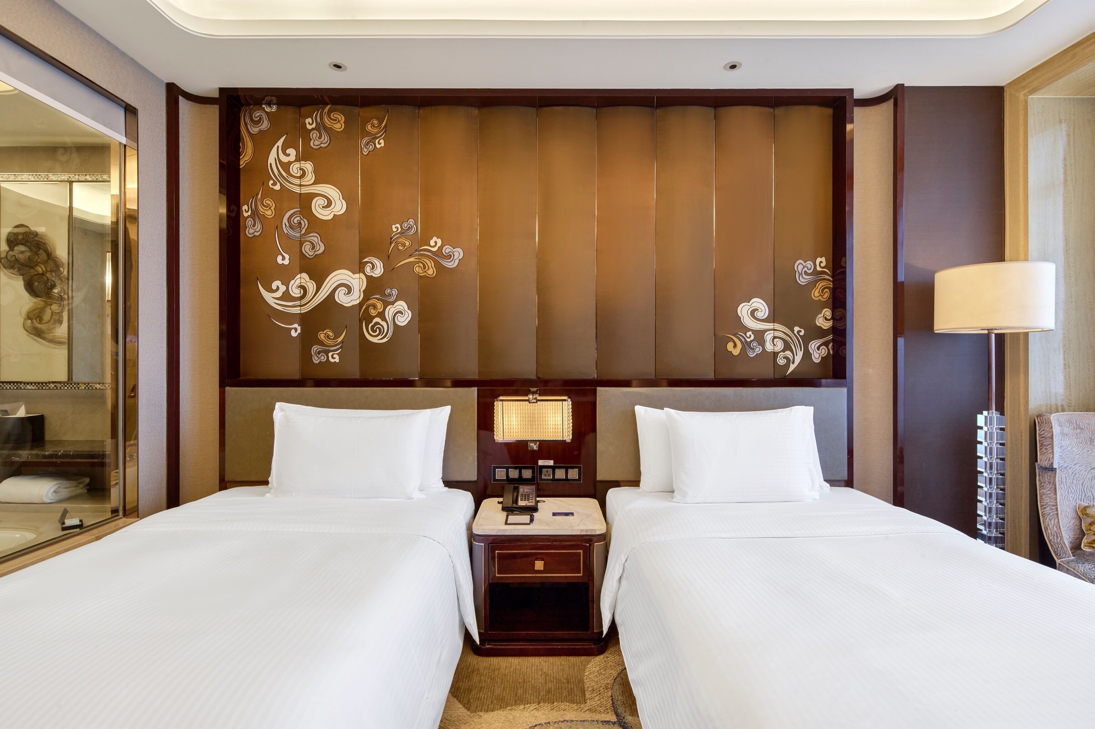 Habitación de hotel con dos camas, cabecero de madera con diseños decorativos, mesita de noche con lámpara y un espejo.
