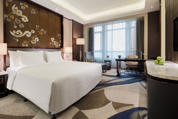 Quarto de hotel com uma cama grande, uma escrivaninha com cadeira, uma mesinha de apoio com abajur e uma janela ampla com cortinas.