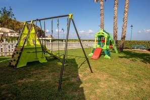 A playground with swings and slides, surrounded by a white picket fence and palm trees. - Arcomagno Hotel Village Club (San Nicola Arcella)