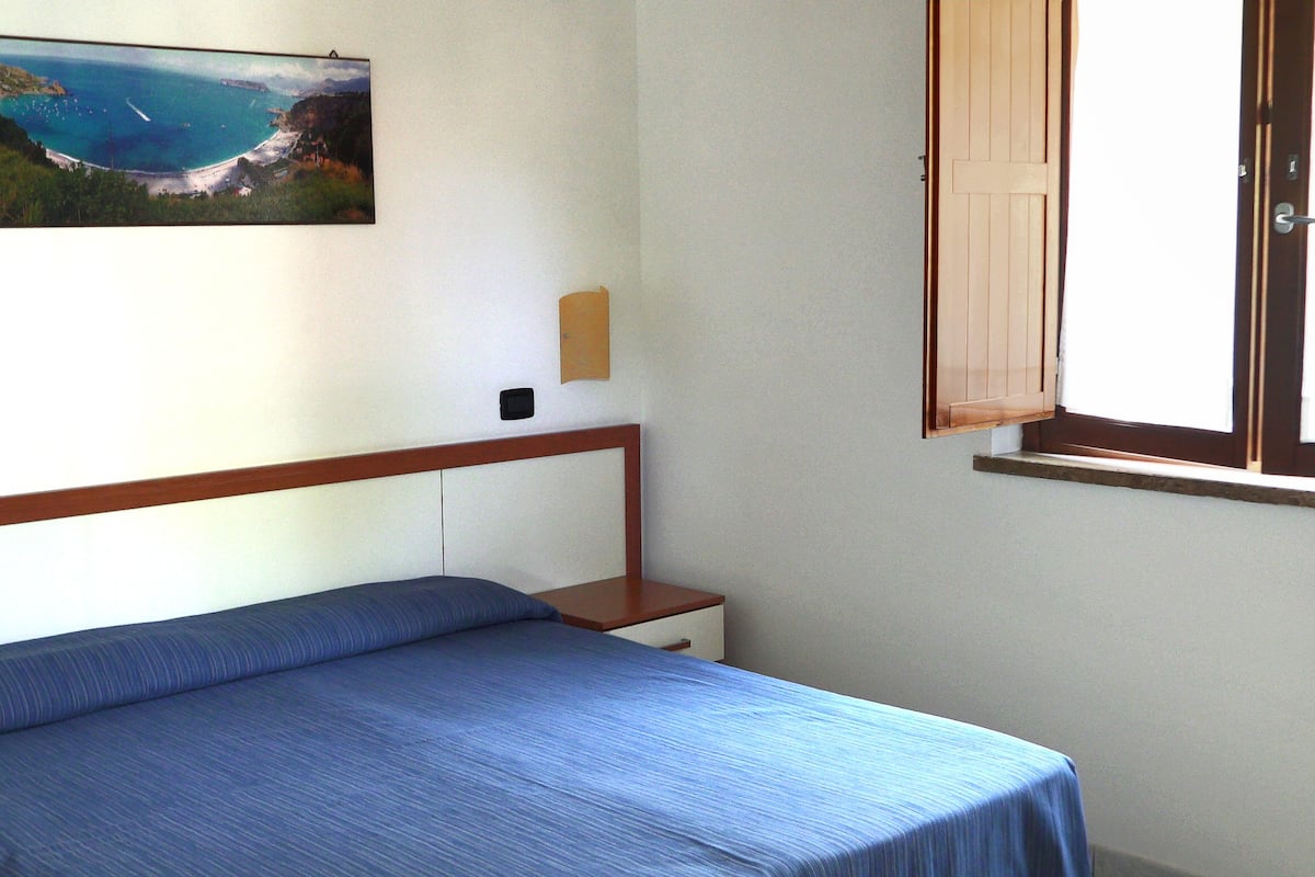 Un dormitorio con una cama, una ventana con contraventanas de madera, un cuadro colgado en la pared que muestra una escena costera y una mesita de noche pequeña.