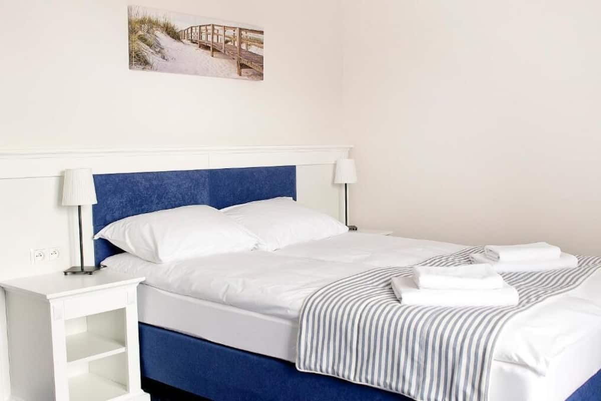 A neatly made bed with white and striped bedding, a blue headboard, and a bedside table with a lamp.
