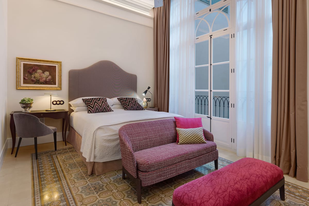 Ein Hotelzimmer mit einem großen Bett, einem Schreibtisch mit Lampe, einem Sessel, einer Bank und einem Fenster mit Vorhängen.