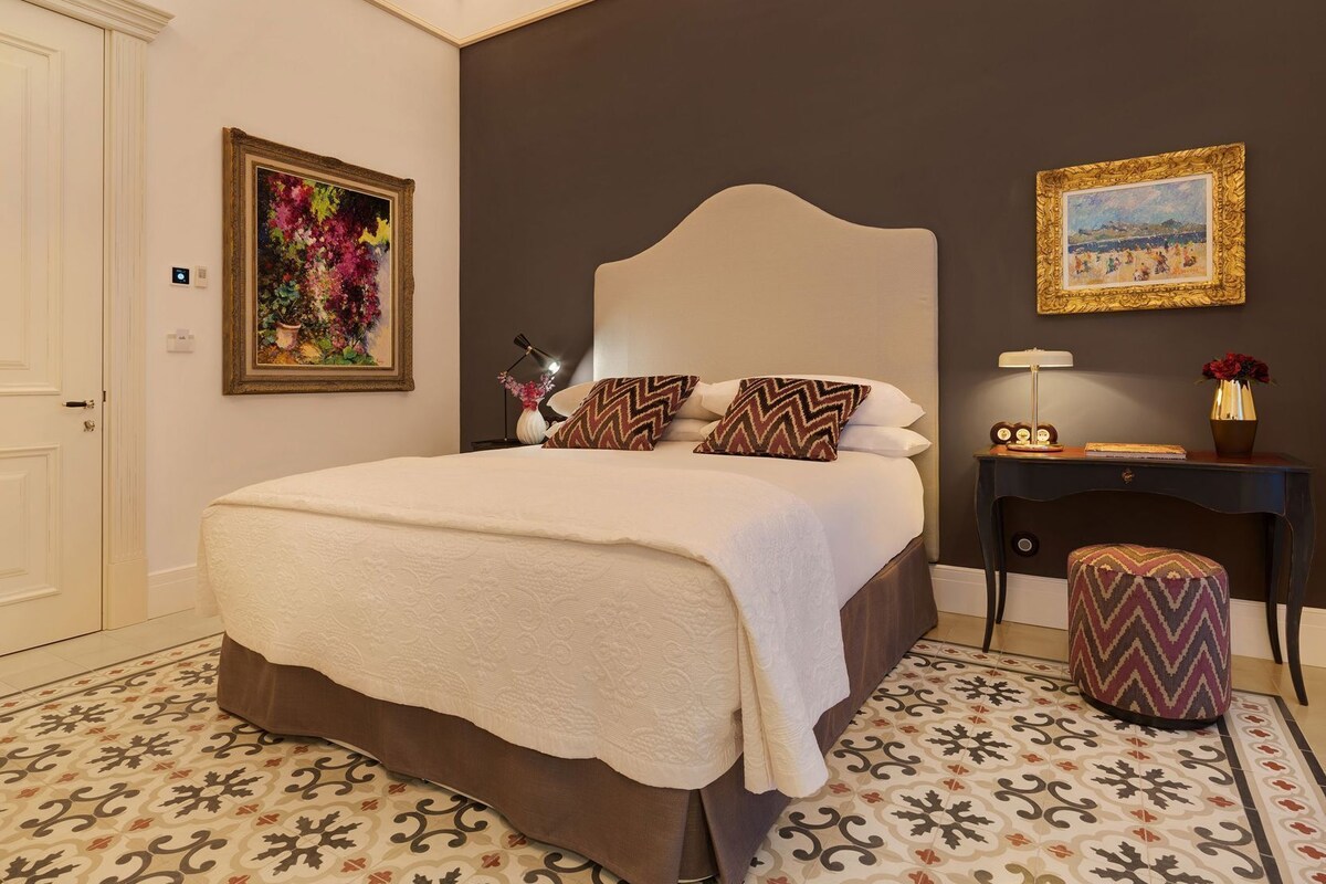 Une chambre à coucher avec un grand lit, une table de chevet, une commode, une chaise et un mur orné de deux tableaux encadrés.