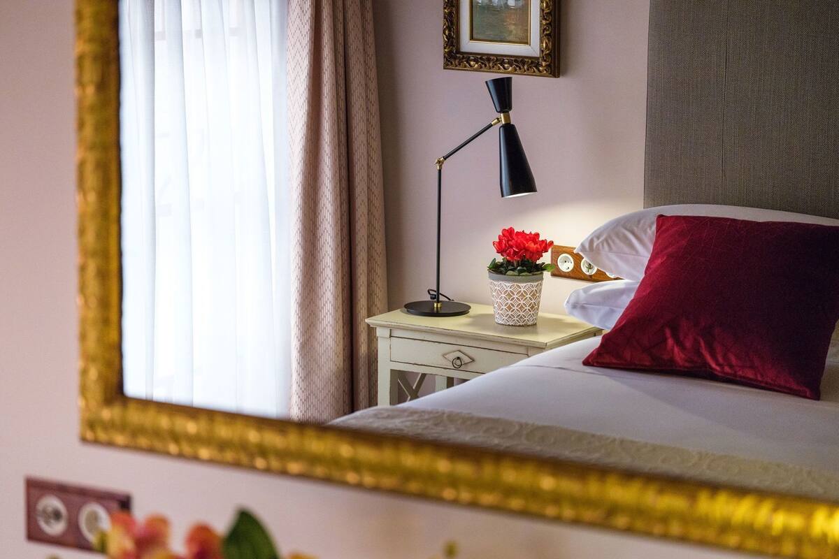 Ein ordentlich bezogenes Bett mit einem roten Kissen, einer Nachttischlampe und einer dekorativen Vase mit Blumen auf einem Nachttisch.