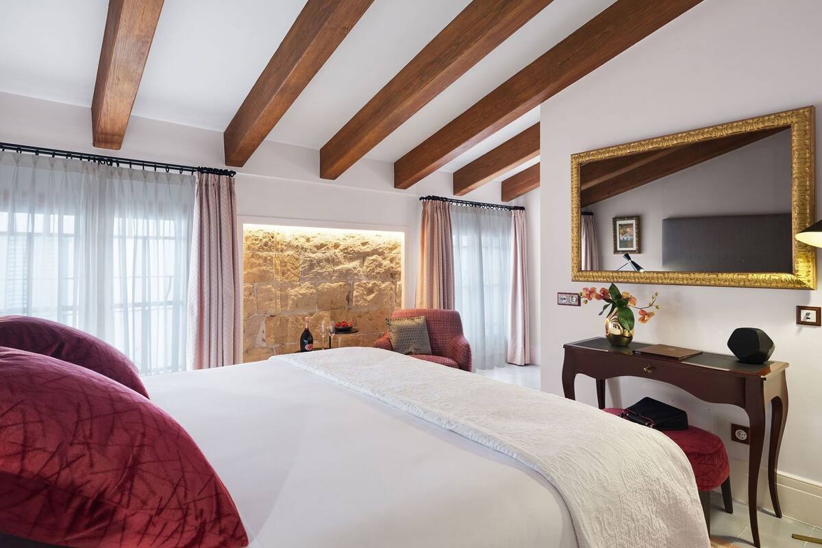Ein Schlafzimmer mit einem großen Bett, Holzbalken, einer Steinwand, einem Spiegel und einem Schreibtisch mit Lampe.