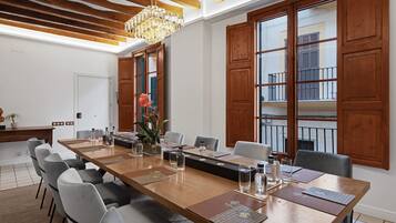 Un salón de conferencias con una larga mesa de madera, sillas grises, una araña y ventanas grandes con contraventanas de madera.