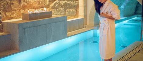 Un área de spa con pared de piedra, piscina climatizada y una persona de bata junto a la piscina.