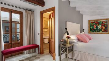 Um quarto com uma cama, um banquinho, uma mesa de cabeceira com luminária, um quadro e vista para o banheiro por uma porta aberta.