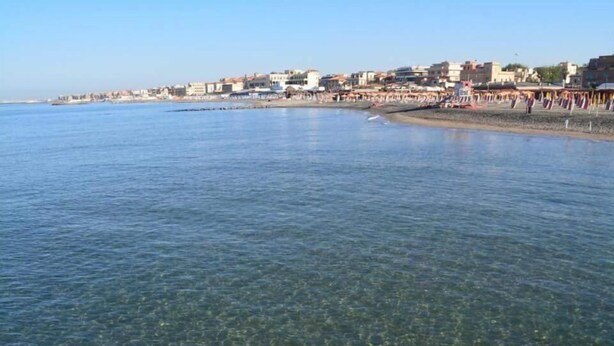 A coastal town with clear waters, buildings along the shore, and a beach with people and umbrellas. - Il Mare di Roma (Rome)