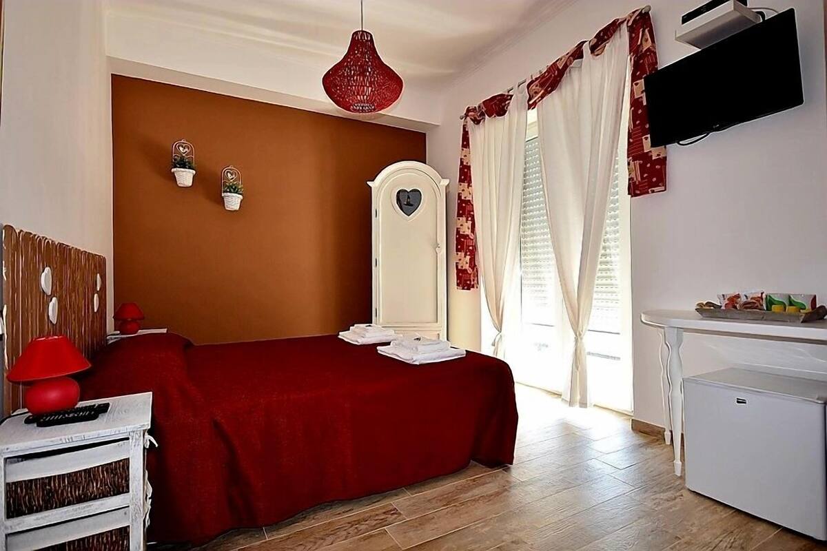 Ein Schlafzimmer mit rotem Bettbezug, weißem Schminktisch, roter Lampe, einem weißen, an der Wand befestigten Fernseher und einem kleinen weißen Kühlschrank.