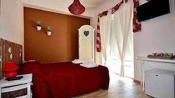 Quarto com colcha vermelha, cómoda branca, luminária vermelha, TV branca fixada na parede e frigorífico pequeno e branco.