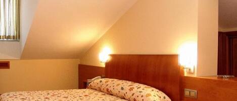 Una habitación de hotel con techo inclinado, una cama con ropa de cama floral, una mesita de noche y un velador.