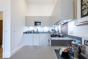 A modern kitchen with white cabinetry, a stainless steel microwave, and a built-in oven. There is a fruit bowl on the counter and a coffee maker. - Lak Serviced Apartments (London)
