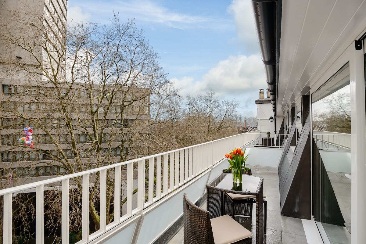 Un balcon agrémenté de meubles en osier, un vase rempli de fleurs et une vue sur un immeuble et des arbres sans feuilles.
