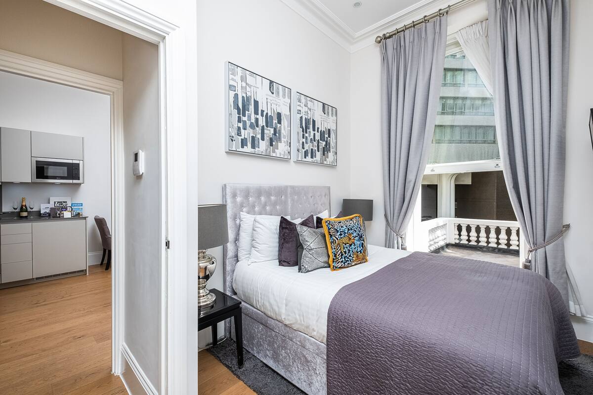 Une chambre comprenant un lit, une table de chevet, une fenêtre avec des rideaux, un balcon et une kitchenette située dans la pièce adjacente.