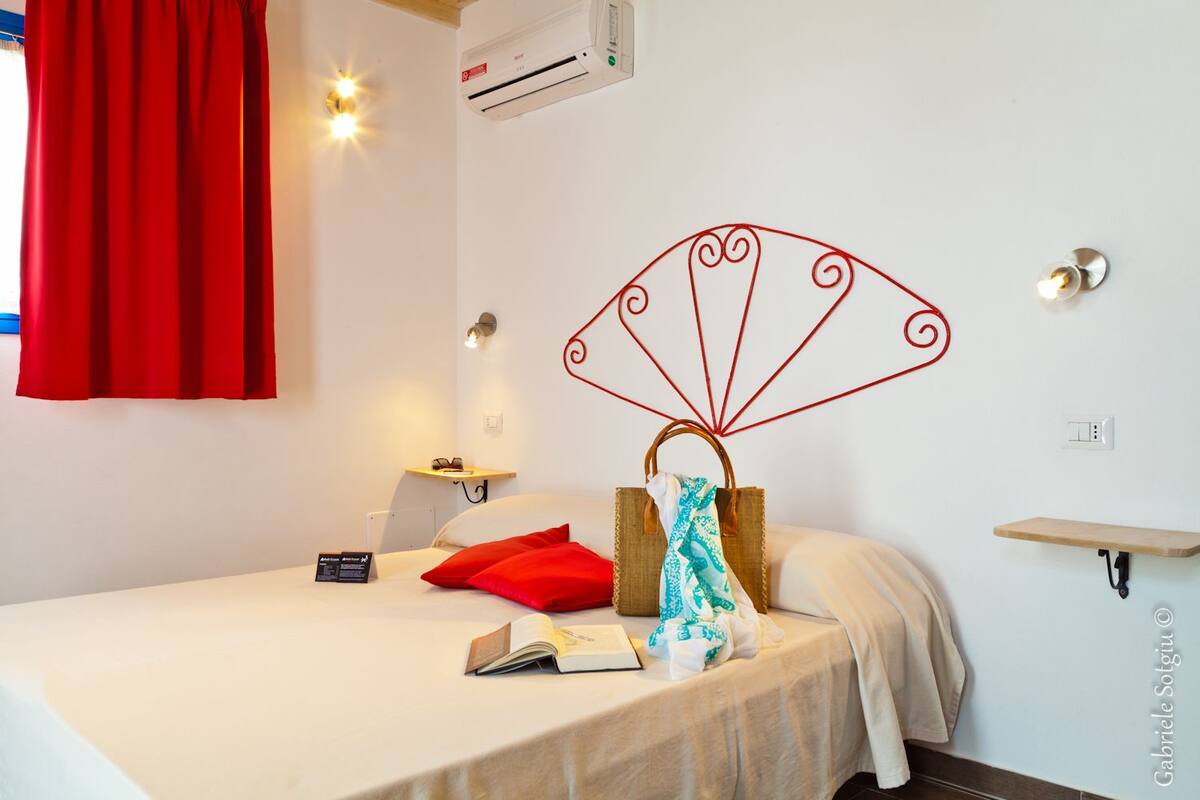 A hotel room with a bed, a red curtain, a wall-mounted air conditioner, a bedside table with a lamp, and a decorative wall piece.