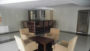 Un área de estar moderna con una mesa central rodeada por cuatro sillones, una zona de bar con varias opciones de asientos, y una pared con diseño.