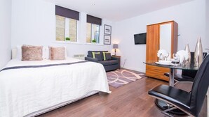 A modern hotel room with a large bed, a sofa, a dining table with chairs, a mirror, and a TV mounted on the wall. - One Whites Row (London)