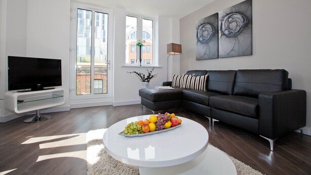 A modern living room with a flat-screen TV, a black leather sofa, a white coffee table with fruit, and large windows with a view of buildings. - One Whites Row (London)