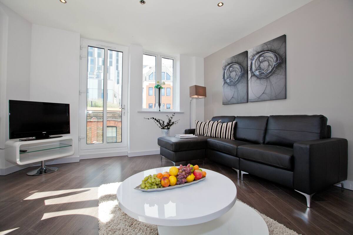 Ein modernes Wohnzimmer mit Fernseher, schwarzem Ledersofa, weißem Couchtisch mit Obst und großen Fenstern mit Blick auf Gebäude.