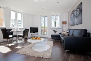 A modern living room with a black leather sofa, a glass coffee table with fruit, a TV, and a large window with a view of buildings.