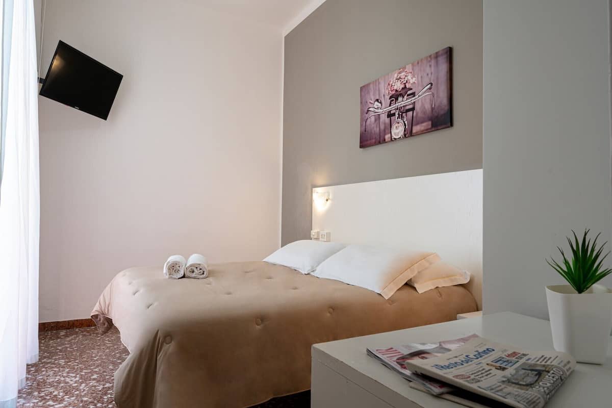 A hotel room with a bed, a flat-screen TV mounted on the wall, a painting above the bed, a bedside table with a newspaper, and a potted plant.