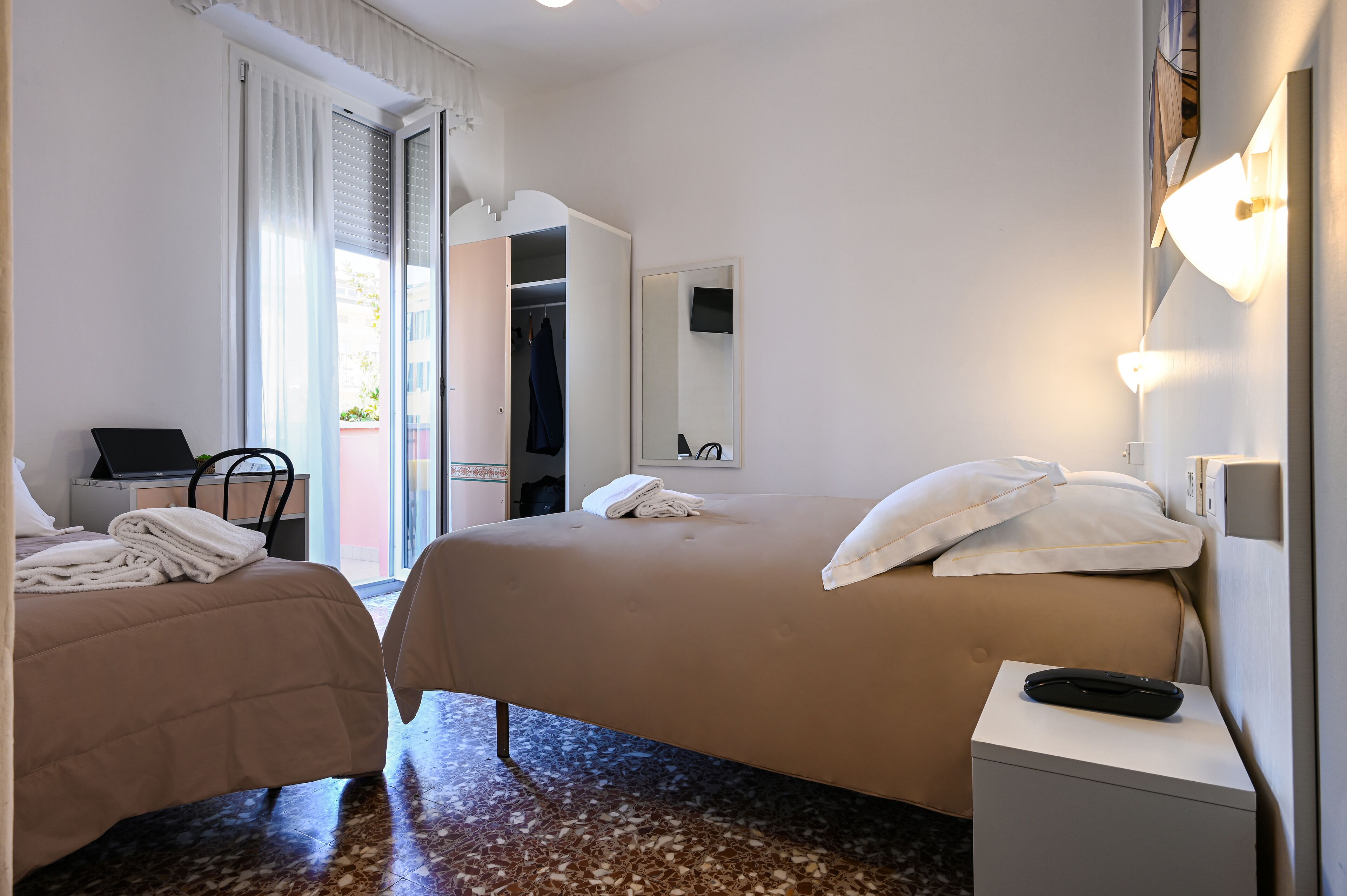 A hotel room with two beds, a desk with a laptop, a chair, and a bathroom with a mirror.