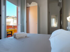 Habitación de hotel con una cama grande, un balcón con mesa y sillas, y una puerta que da a otra habitación.
