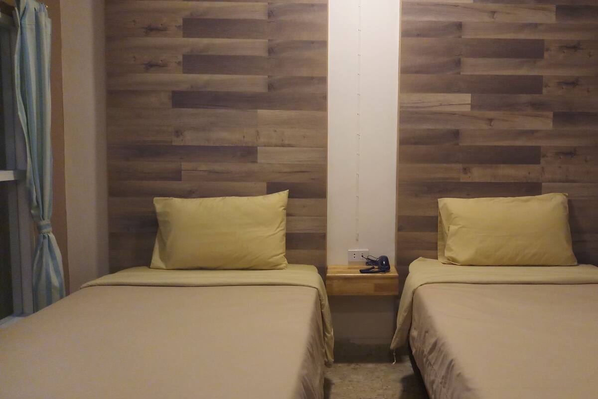 A room with two beds, each with a pillow and a bedside table with a lamp.