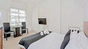 A hotel room with a bed, a desk, chairs, a TV, and a window with blinds.