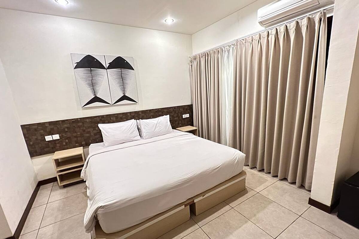 A modern hotel room with a large bed, a headboard with a geometric design, a wall-mounted air conditioner, and a window with curtains.