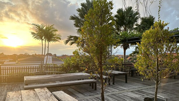 A wooden deck with benches and potted trees, overlooking a body of water and buildings. - The Paving Villa (Canggu)