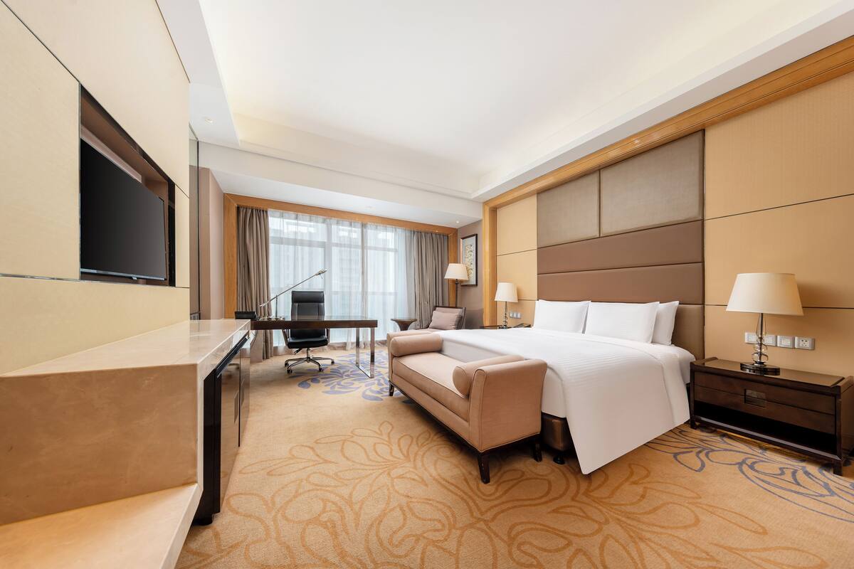 A hotel room with a bed, desk, chair, TV, and lamps.