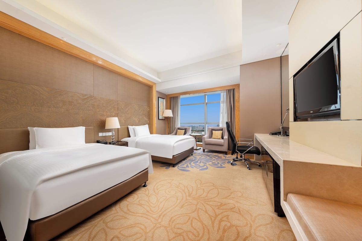 A hotel room with two beds, a desk, a TV, and a view of the city.