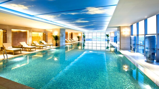 Una piscina interior moderna con agua azul cristalina, sillas de descanso y vistas al cielo a través del techo.