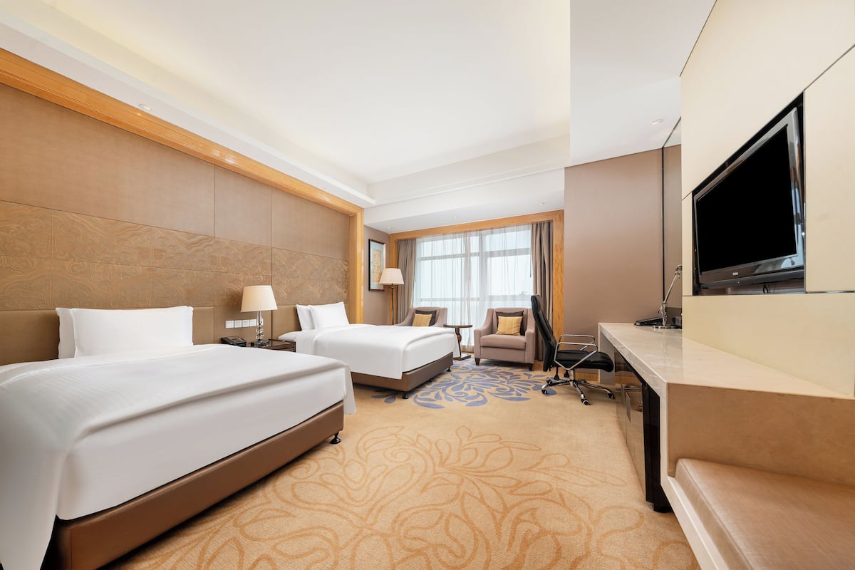 A hotel room with two beds, a desk with a flat-screen TV, a chair, and a window with curtains.