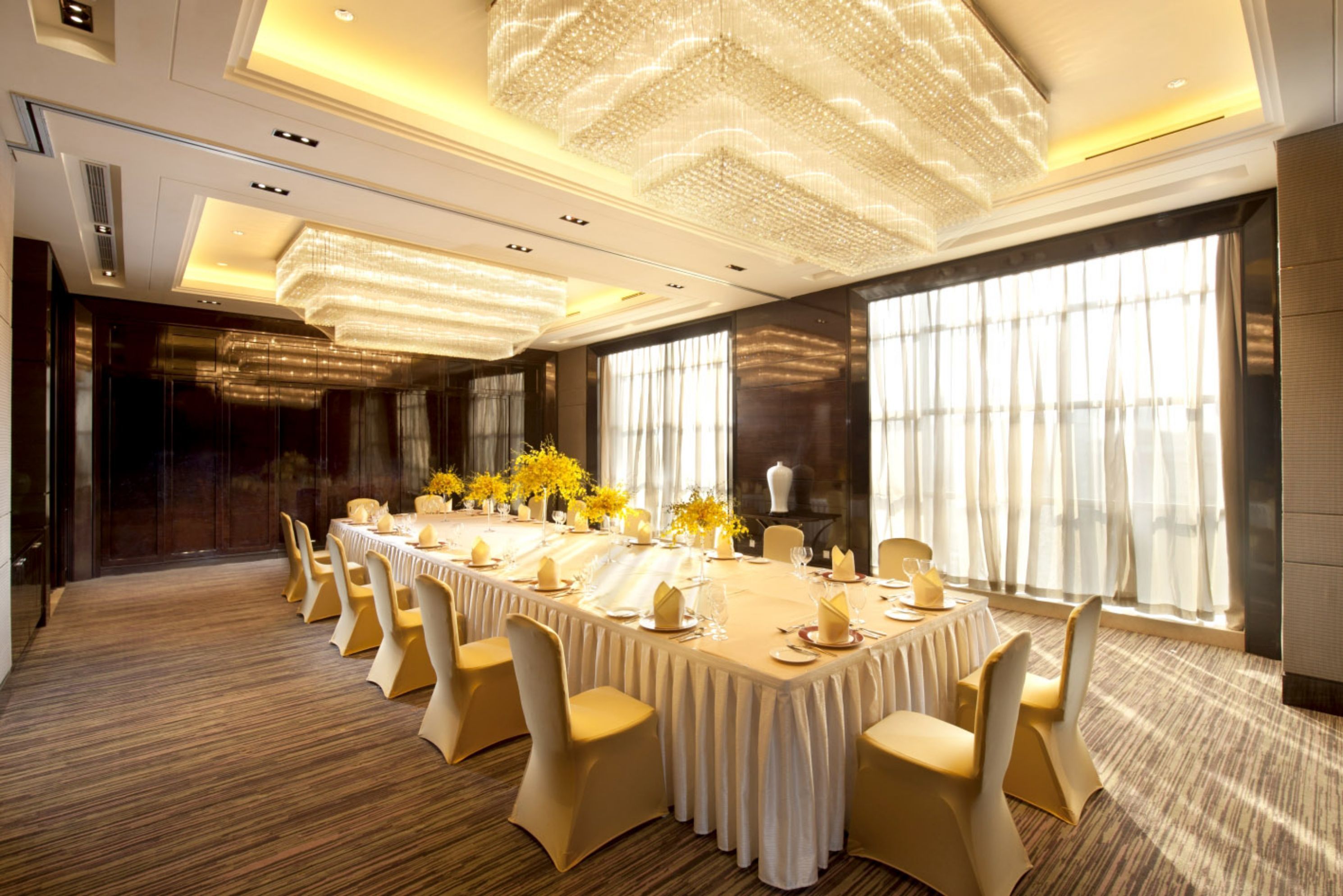 Un salón de banquetes con una larga mesa dispuesta para un evento formal, rodeado de sillas, grandes ventanales y una araña.