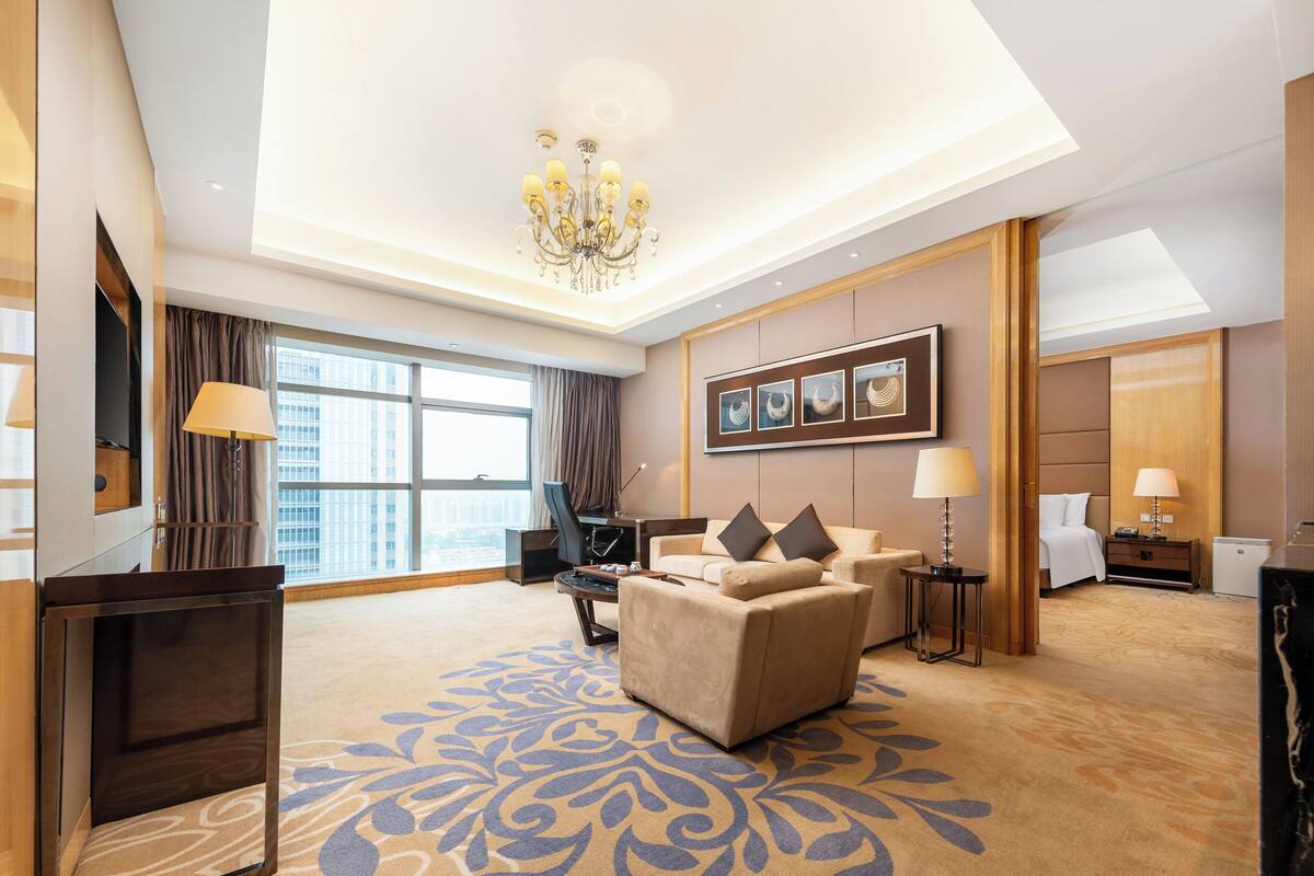 A hotel room with a large window, a chandelier, a sofa, a desk, and a bed.