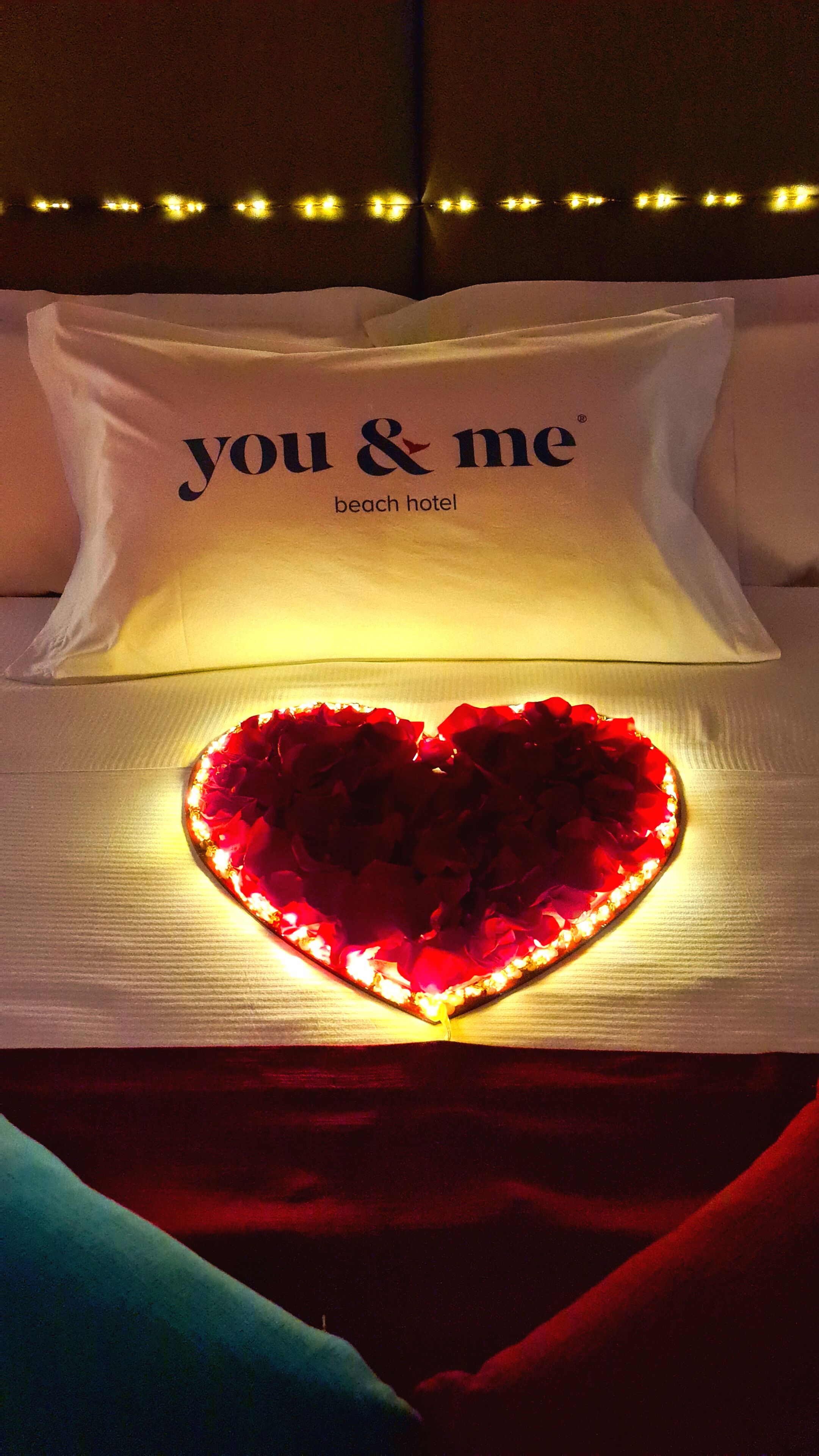 Una cama con un almohadón que dice "you & me beach hotel" y una lámpara decorativa con forma de corazón sobre la cama.