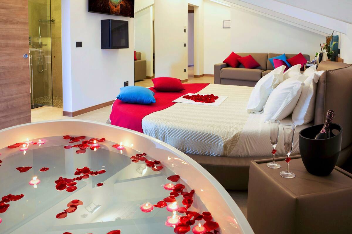 Ein modernes Hotelzimmer mit einem großen Bett, einer Sitzecke mit einer Couch, einer gläsernen Dusche und einer Whirlwanne mit Rosenblättern und Kerzen.