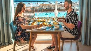 A dining table with plates of food, a man and a woman seated, and a view of the sea and beach through large windows. - You & Me Beach Hotel (Rimini)