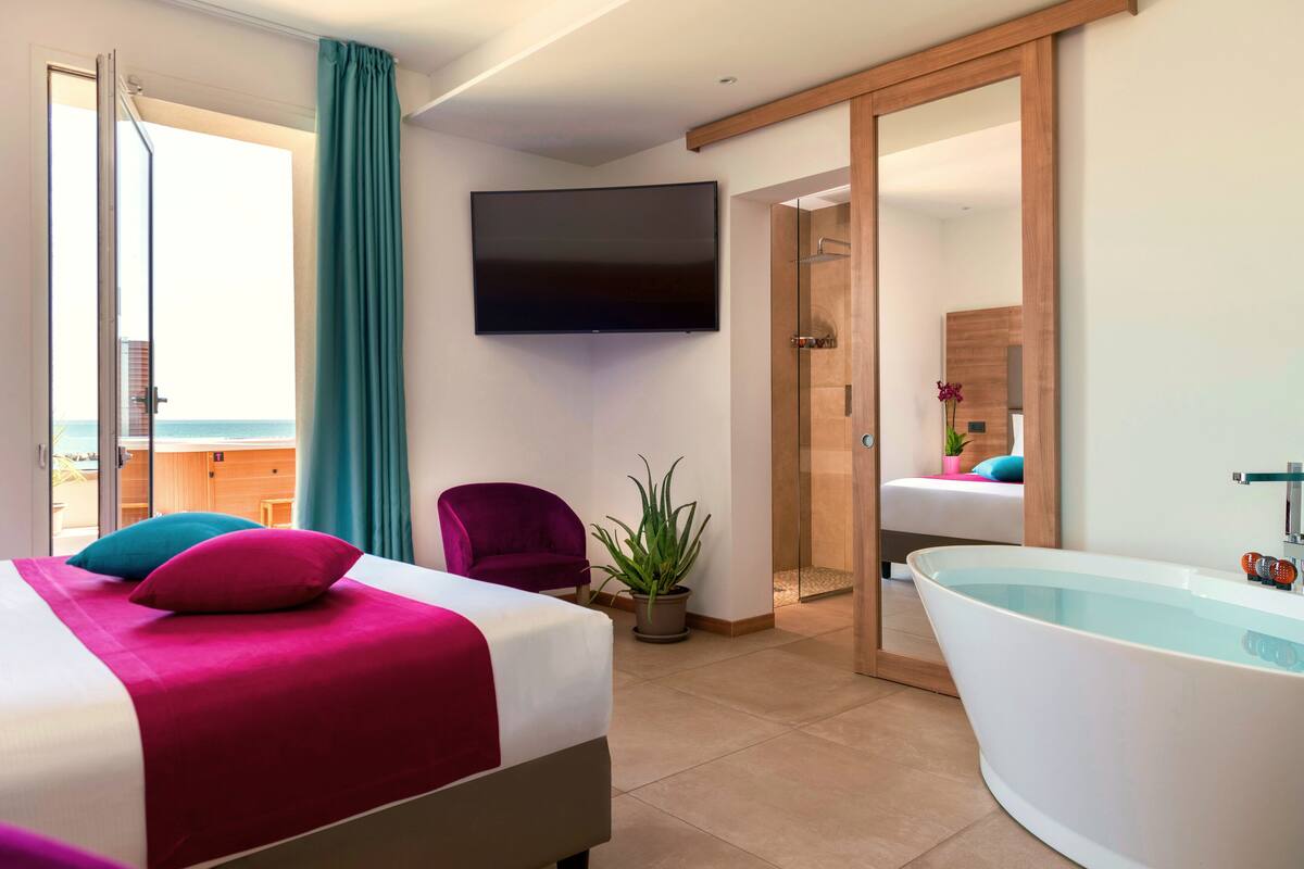 Ein modernes Hotelzimmer mit einem Bett, einem großen Flachbildfernseher, einer Badewanne und einer Glastür, die auf einen Balkon führt.