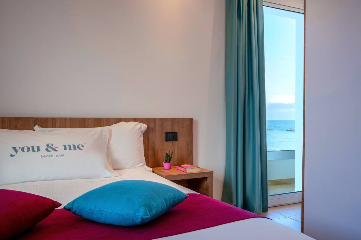 Ein Hotelzimmer mit Bett, Kissen, einem Nachttisch, einer Zimmerpflanze und Meerblick.