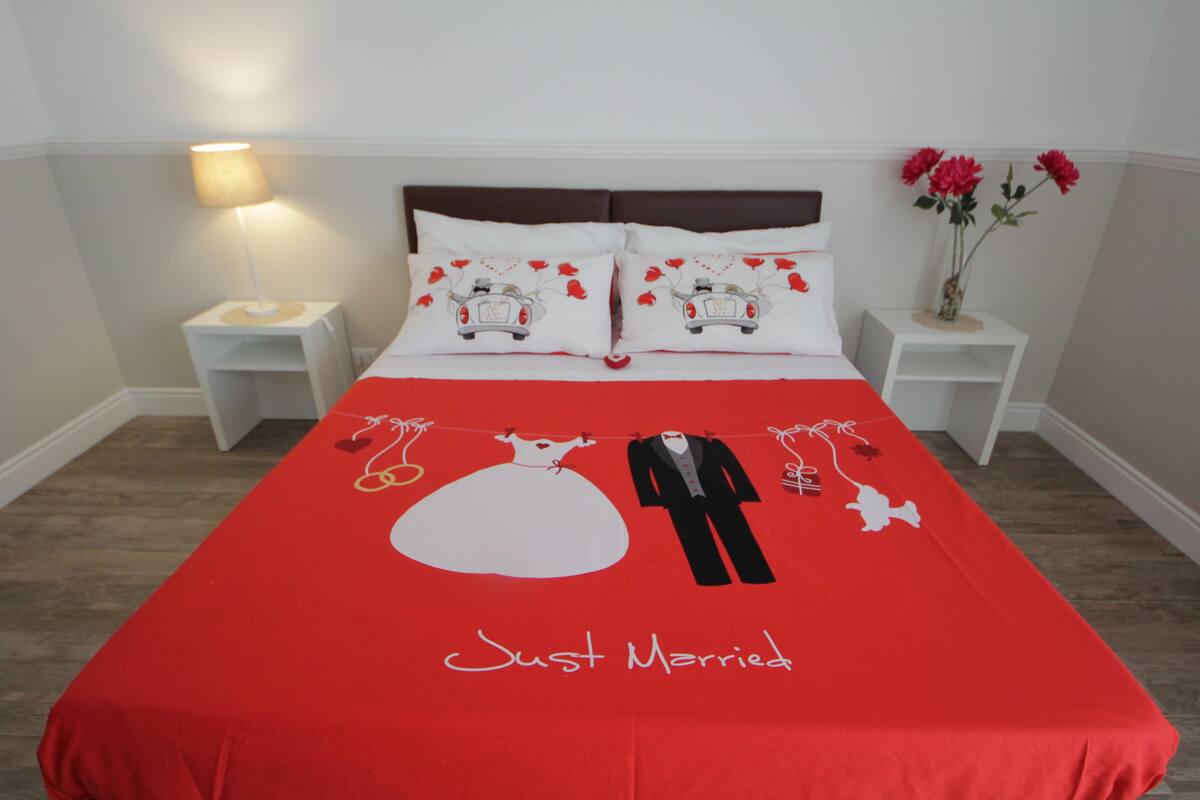 A hotel room with a red bedspread featuring a wedding theme, white pillows, a nightstand with a lamp, and a vase with flowers on the side table.