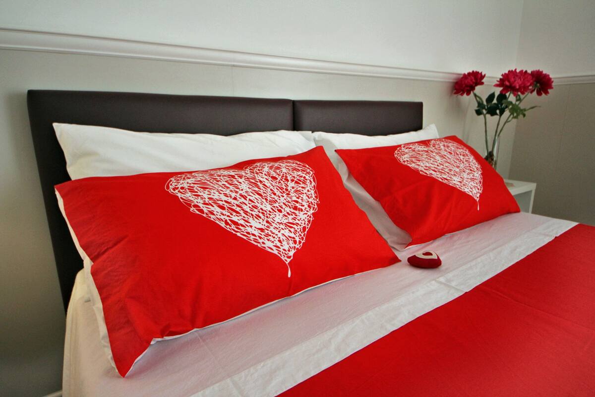 A bed with red and white bedding, a dark headboard, and a vase with red flowers on a nightstand.