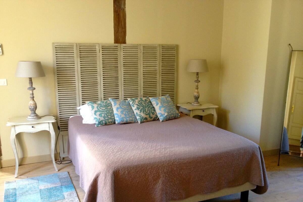 A bedroom with a bed, two bedside tables, a nightstand, a window with shutters, and a door.