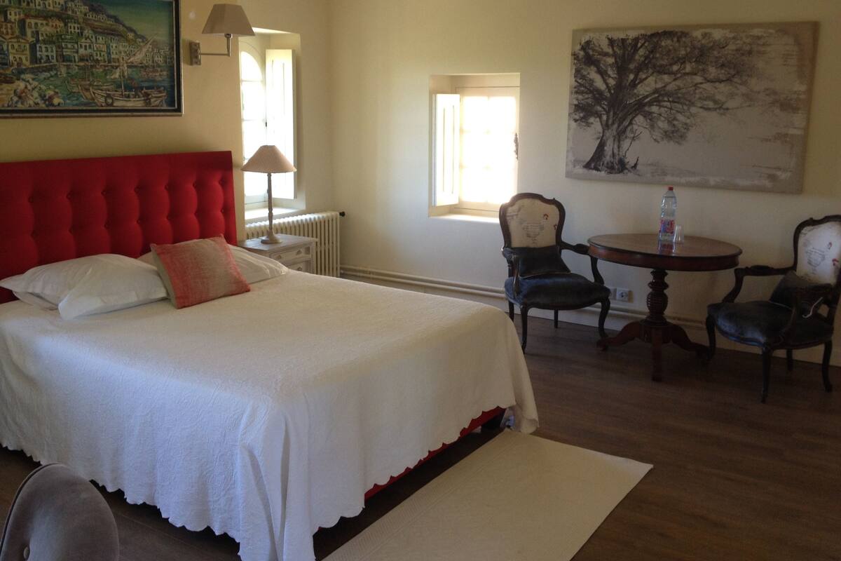 A hotel room with a large bed, a dining table with chairs, a radiator, and two paintings on the walls.