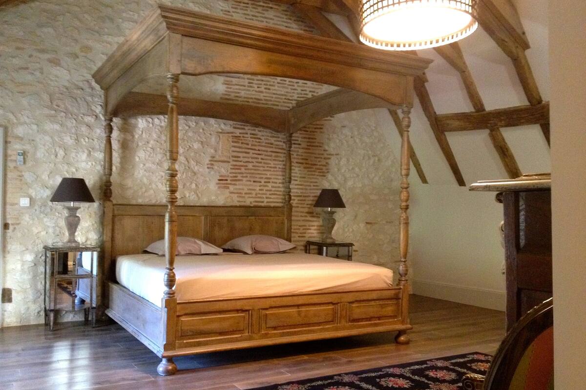A four-poster bed in a room with stone walls and wooden beams. There are two bedside tables with lamps.