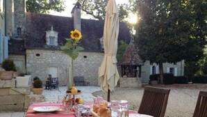 Mesa de comedor al aire libre con platos, vasos y una cesta de pan, con un edificio de piedra y árboles al fondo.
